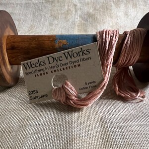 May include: A wooden spool with pink cotton floss. The spool has dark brown ends and a light brown center with a blue stripe. A tag reads "Weeks Dye Works" and "Sanguine" with 5 yards of cotton floss.