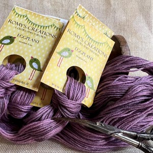 May include: Purple embroidery floss wound around a spool with a label that reads "Romys Creations Colors and Threads Eggplant".