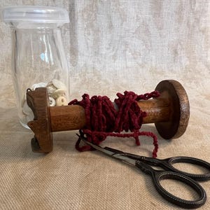 May include: A wooden spool with a red yarn wrapped around it, a glass jar filled with white buttons, and a pair of silver scissors on a brown fabric surface.