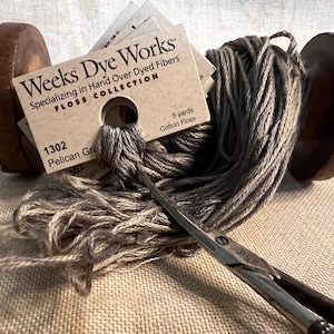 May include: A spool of Weeks Dye Works Pelican Gray cotton floss, color number 1302, with 5 yards of floss. The spool is on a wooden stand and a pair of scissors is lying on top of the floss.
