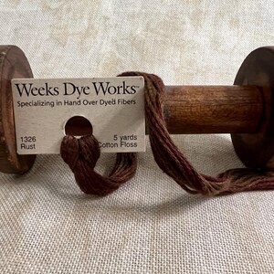 May include: A wooden spool of Weeks Dye Works hand-dyed cotton floss in the color Rust (1326). The floss is a blend of brown, red, and beige shades. The spool is 5 yards of cotton floss.