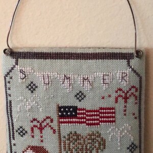 May include: A hand-stitched, square wall hanging with a patriotic theme. The design features an American flag, palm trees, houses, and the word "SUMMER" in banner letters. A metal wire is attached for hanging.