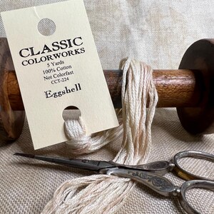 May include: A spool of Eggshell colored thread, 100% cotton, 5 yards, not colorfast, on a wooden spool. The thread is wound around the spool and is ready to be used for sewing or embroidery. The label reads "Classic Colorworks, 5 Yards, 100% Cotton, Not Colorfast, CCT-224, Eggshell".