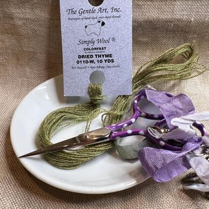 May include: A white card with the text "The Gentle Art, Inc. Purveyors of hand-dyed, over-dyed threads Simply Wool® COLORFAST by industry standards DRIED THYME 0110-W, 10 YDS P.O. Box 670 • New Albany, Ohio 43054"  A skein of green yarn is threaded through a hole in the card. A pair of purple scissors with white polka dots is on a white plate with a skein of green yarn, a purple ribbon, and a white ribbon.