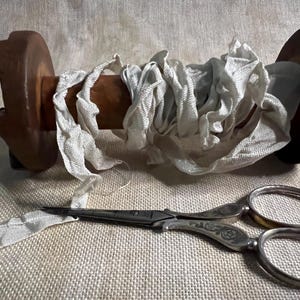 May include: A spool of white fabric ribbon wrapped around a wooden spool with a pair of silver scissors with ornate handles.