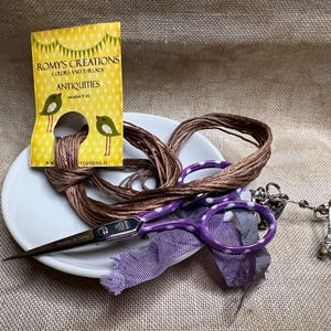 May include: A white ceramic dish with a brown embroidery floss, purple scissors with white polka dots, and a silver charm with a key and Eiffel Tower design. The dish also has a yellow card with the text "Romy's Creations Colors and Threads Antiques".