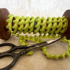 May include: A spool of lime green pom-pom trim wrapped around a vintage wooden bobbin, accompanied by a pair of dark metal sewing scissors. The trim features a continuous row of small, soft, spherical pom-poms attached to a woven fabric tape.