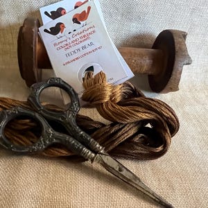May include: A still-life image featuring a wooden thread spool, a skein of brown embroidery floss, and ornate scissors. A tag reads "Romy's Creations COLORS AND THREADS TEDDY BEAR". The scissors are silver with decorative handles. The floss is a warm, earthy brown.