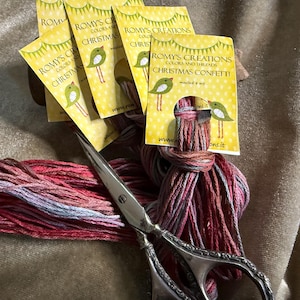May include: A bundle of colorful embroidery floss with scissors and several yellow tags. The tags read "ROMY'S CREATIONS COLORS AND THREADS CHRISTMAS CONFETTI" and feature a bird illustration. The floss is red, pink, and gray.
