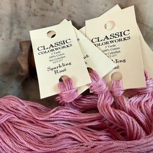 May include: Close-up of pink embroidery floss labeled "Sparkling Rosé" by Classic Colorworks. Each skein is 5 yards of 100% cotton, non-colorfast. The floss is a soft, light pink color, perfect for embroidery and other crafts.