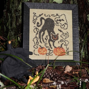 May include: A black cat with a white face and a black tail is standing on its hind legs. The cat is surrounded by vines and pumpkins. The image is in a black frame.
