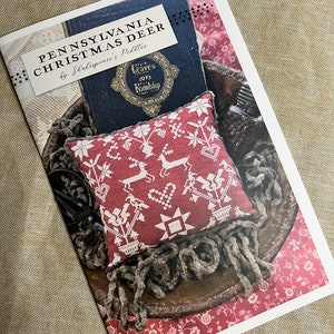 May include: A printed pattern for a cross-stitch project called "Pennsylvania Christmas Deer" by Shakespeare's Peddler. The pattern features a red and white deer with a heart and a star design. The pattern is displayed on a book with a brown wooden bowl and a brown fuzzy rope.