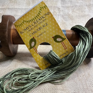 May include: A spool of green embroidery floss with a yellow label that reads "Romy's Creations Colors and Threads Camouflage mouliné 8 cct www.romyscreations.it".