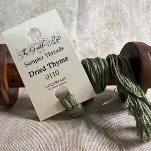 May include: A spool of green thread labeled "The Gentle Art Sampler Threads Dried Thyme 0110 COLORFAST" on a wooden spool.