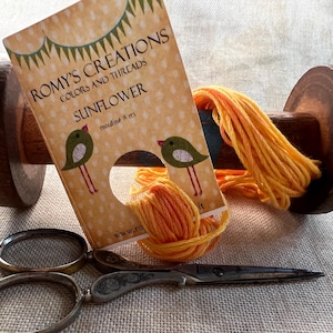 May include: A wooden thread spool with bright orange thread, a card of "Sunflower" embroidery floss, and small silver scissors. The card features two green birds with red legs. The text "Romy's Creations" is visible.
