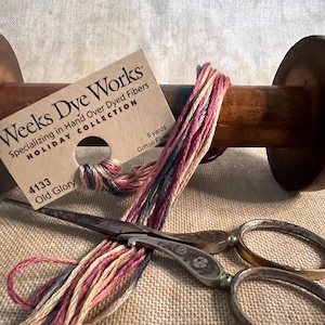 May include: A spool of Weeks Dye Works "Old Glory" hand-dyed cotton embroidery floss in shades of red, white, and blue. The floss is wrapped around a wooden spool and is accompanied by a label with the company name, product name, and color number. The floss is also accompanied by a pair of antique scissors.