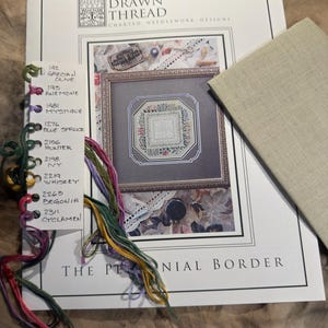 May include: A cross-stitch pattern booklet titled The Drawn Thread Charted Needlework Designs The Perennial Border, featuring a color-coded thread organizer card with labeled embroidery floss in various shades and a piece of beige linen fabric.