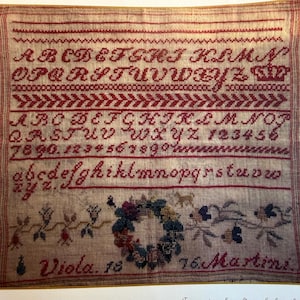 May include: A framed antique sampler with red cross-stitch lettering and decorative borders. The sampler includes the alphabet, numbers, and the names "Viola. 18" and "76. Martini". The bottom features a floral wreath.