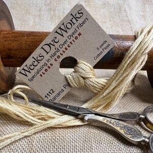 May include: A skein of hand-dyed cotton floss in a pale yellow color, labeled "Weeks Dye Works" and "1112 Banana Pu". The floss is wrapped around a wooden spool and a pair of small scissors are lying on top of the floss.