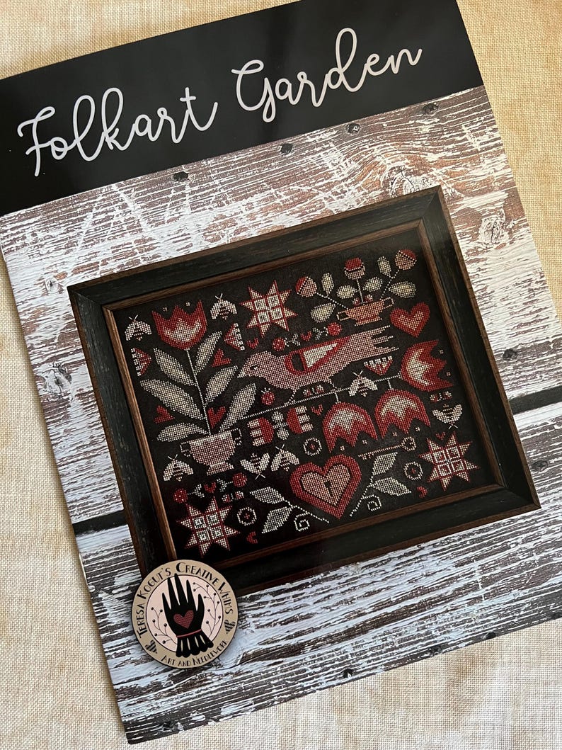 May include: Folkart Garden cross stitch pattern featuring a framed design with a red heart, birds, flowers, and stars. The pattern is by Teresa Kogut's Creative Stitches.