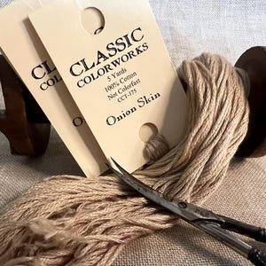 May include: A skein of beige embroidery floss wound on a wooden spool, with two cards labeled "Classic Colorworks" and "Onion Skin". The cards indicate the floss is 5 yards of 100% cotton, not colorfast, and has the color code CCT-175.