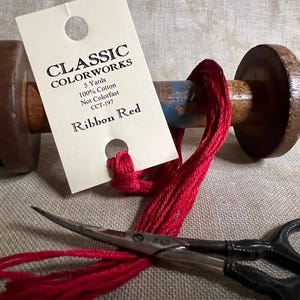 May include: A spool of red ribbon yarn with a label that reads "Classic Colorworks 5 Yards 100% Cotton Not Colorfast CCT-197 Ribbon Red".