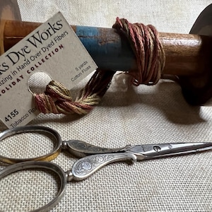 May include: A wooden spool of hand-dyed cotton floss in shades of red, brown, and gold. The spool is labeled "Weeks Dye Works" and "Holiday Collection". The floss is 5 yards long. A pair of antique silver scissors with ornate handles are also pictured.