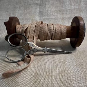 May include: A spool of beige twill tape wrapped around a wooden spool with a pair of antique silver scissors resting on top.