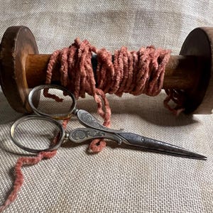 May include: A wooden spool of pink yarn with a pair of ornate silver scissors. The spool is made of wood and has a decorative design. The scissors have a floral design on the handles.