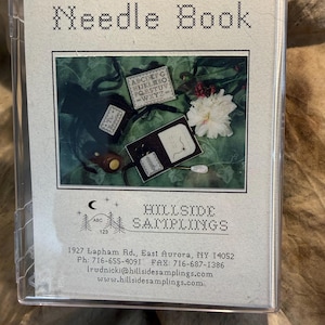 May include: A clear plastic case containing a craft kit labeled Alphabet Needle Book by Hillside Samplings. The insert features a photograph of finished needlework projects, including a small alphabet sampler, a needle book, and sewing tools.