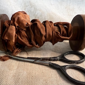 May include: A spool of brown ribbon wrapped around a wooden spool with a pair of antique silver scissors.