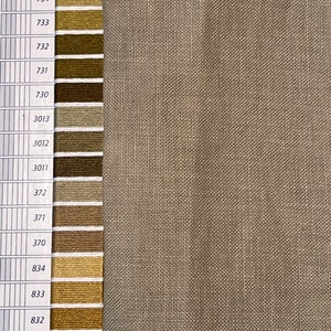 May include: A large piece of light brown, textured fabric positioned next to a vertical color chart. The chart displays a column of numbered color swatches ranging from olive green to dark brown, including numbers 581, 580, 734, 733, 732, 731, 730, 3013, 3012, 3011, 372, 371, 370, 834, 833, 832, 831, and 830.