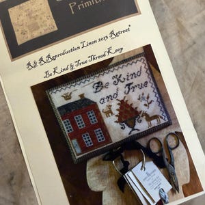 May include: A printed pattern booklet titled Stacy Nash Primitives featuring a cross-stitch design titled Be Kind and True. The design shows a red house, a tree, and animals. Included are metal scissors and a ring with small paper tags attached.