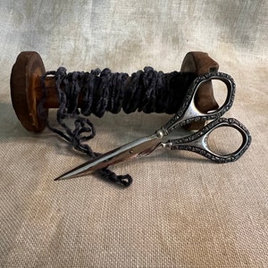 May include: A pair of silver-plated scissors with an ornate design, lying on a spool of dark gray thread.