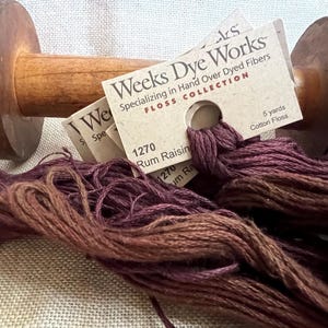May include: A wooden thread spool with a dark brown finish, holding a bundle of variegated floss in shades of purple and brown. The floss is labeled "Rum Raisin" and "Weeks Dye Works".