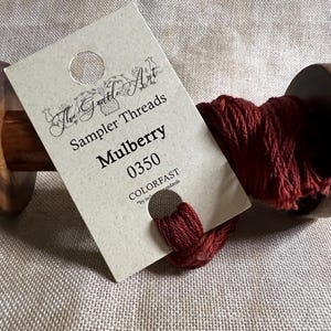 May include: A spool of mulberry colored thread wound on a wooden bobbin. The thread is labeled "The Gentle Art Sampler Threads Mulberry 0350 COLORFAST"