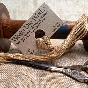 May include: A spool of tan cotton floss wound around a wooden bobbin. The floss is labeled "Weeks Dye Works" and "1121 Straw" with a 5-yard length. A pair of antique silver scissors are also pictured.