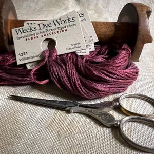 May include: A spool of burgundy embroidery floss wound around a wooden bobbin. The floss is labeled "Weeks Dye Works" and "Williamsburg 1321". A pair of antique silver embroidery scissors are also pictured.