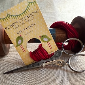May include: A wooden thread spool with red thread, a pair of ornate scissors, and a card that reads "ROMY'S CREATIONS Colors and Threads Red Passion". The card features green bird illustrations and the text "mouline 8 m".
