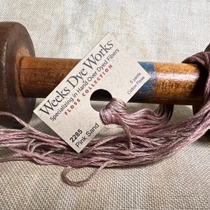 May include: A spool of pink sand colored cotton floss with a label that reads "Weeks Dye Works FLOSS COLLECTION Specializing in Hand Over Dyed Fibers 2285 Pink Sand 5 yards Cotton Floss"