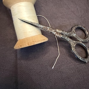 May include: A wooden spool of white thread paired with a pair of silver-toned metal scissors featuring an ornate, embossed handle design. The scissors are positioned diagonally across the spool, with a single strand of white thread extending outward.
