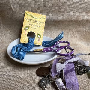 May include: A close-up of a white ceramic dish with blue and purple embroidery floss, a pair of purple scissors with white polka dots, and a charm bracelet with a purple ribbon. The dish is on a beige burlap surface. The embroidery floss is labeled "Romys Creations" and "Simonas Iris".