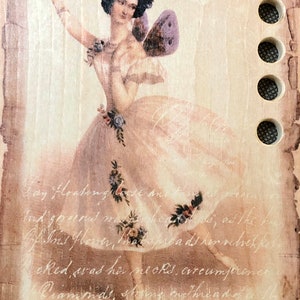 May include: A rectangular wooden plaque featuring a printed image of a dancer with butterfly wings, wearing a light-colored dress adorned with floral accents. The surface includes cursive script text and a vertical row of five circular holes on the right.