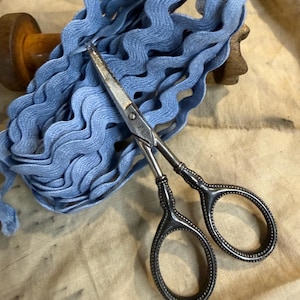 May include: A spool of light blue wavy rickrack trim made of woven fabric, positioned behind a pair of vintage-style metal scissors with ornate, textured handles and the letters P&B engraved on the silver-toned metal blade section.
