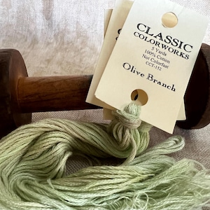 May include: A wooden thread holder with a skein of light green thread and tags that read "Classic Colorworks Olive Branch". The thread is 100% cotton and is 5 yards in length.