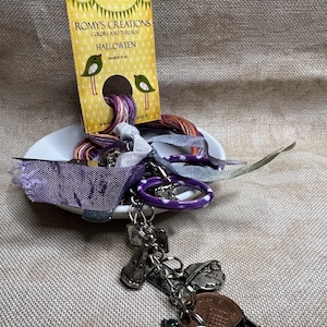 May include: A white ceramic dish with purple, orange, and blue embroidery floss, a white fabric scrap, and a purple and white key chain with charms. The dish is on a beige fabric background. The embroidery floss is labeled "Romys Creations Colors and Threads Halloween".