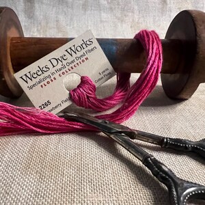 May include: A spool of pink cotton floss with a label that reads "Weeks Dye Works Specializing in Hand Over Dyed Fibers FLOSS COLLECTION 2265 Strawberry Field 5 yards Cotton Floss". The spool is on a wooden spindle with a metal base. A pair of silver embroidery scissors are lying on a linen fabric.