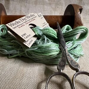 May include: A wooden spool of green and white hand-dyed embroidery floss with two labels that read "Weeks Dye Works" and "2181 Cactus" and "2101 Cactus". A pair of antique silver embroidery scissors are lying on top of the floss.