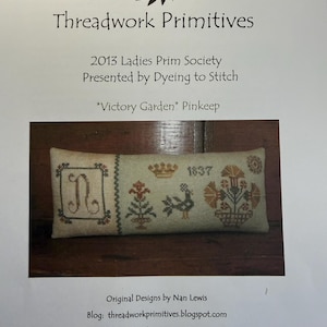 May include: A rectangular cross-stitch pinkeep featuring the text Victory Garden Pinkeep, Threadwork Primitives, 2013 Ladies Prim Society, Presented by Dyeing to Stitch, and Original Designs by Nan Lewis. The embroidery displays a letter N, a crown, a bird, and floral motifs.