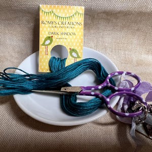 May include: A white ceramic dish with a teal embroidery floss, purple scissors with white polka dots, and a yellow card with the text "ROMY'S CREATIONS COLORS AND THREADS DARK SHADOW" and two green birds.